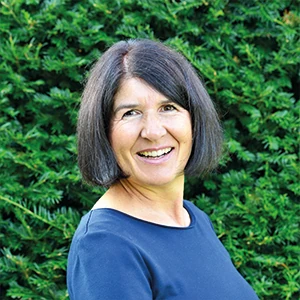 Frau mit dunklen Haaren lächelt vor grüner Hecke, Porträt im Freien bei Tageslicht.