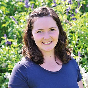 Junge Frau mit braunen lockigen Haaren lächelt im Garten mit grünen Pflanzen und Blumen.