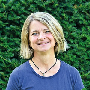 Frau mit blonden Haaren lächelt im Garten vor einer grünen Hecke bei Tageslicht.