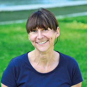 Frau mit braunen Haaren und blauem T-Shirt lächelt im Grünen, Natur im Hintergrund.