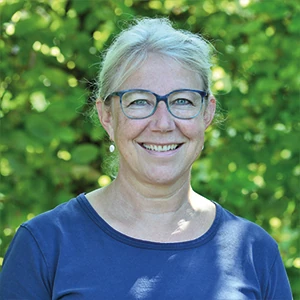 Frau mit Brille und grauen Haaren lächelt im Garten vor grüner Natur im Sommer.