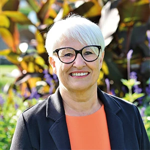 Ältere Frau mit weißem Haar und Brille lächelt im Garten vor grünen Pflanzen.