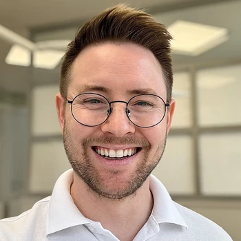 Lächelnder Mann mit Brille und Bart in heller, moderner Büro-Umgebung.