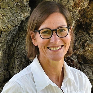 Frau mit Brille und weißer Bluse lächelt vor einem alten, knorrigen Baum im Freien.