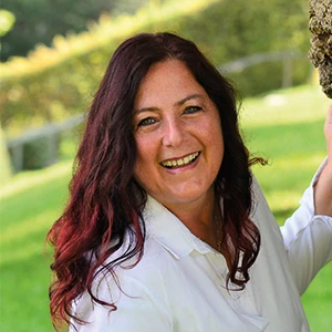 Frau mit langen, dunklen Haaren lächelt in weißer Bluse vor grünem Naturhintergrund.