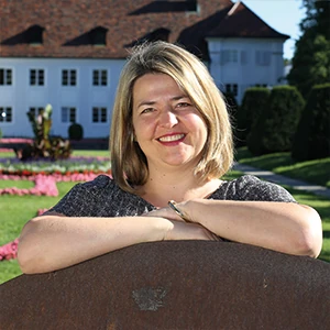 Frau lächelt im sonnigen Schlossgarten, Blumenbeet und historisches Gebäude im Hintergrund.