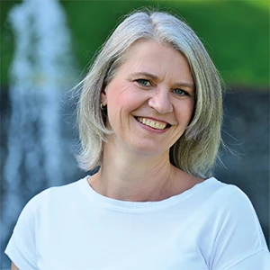 Porträt einer lächelnden Frau mit grauen Haaren im Freien vor grünem Hintergrund.